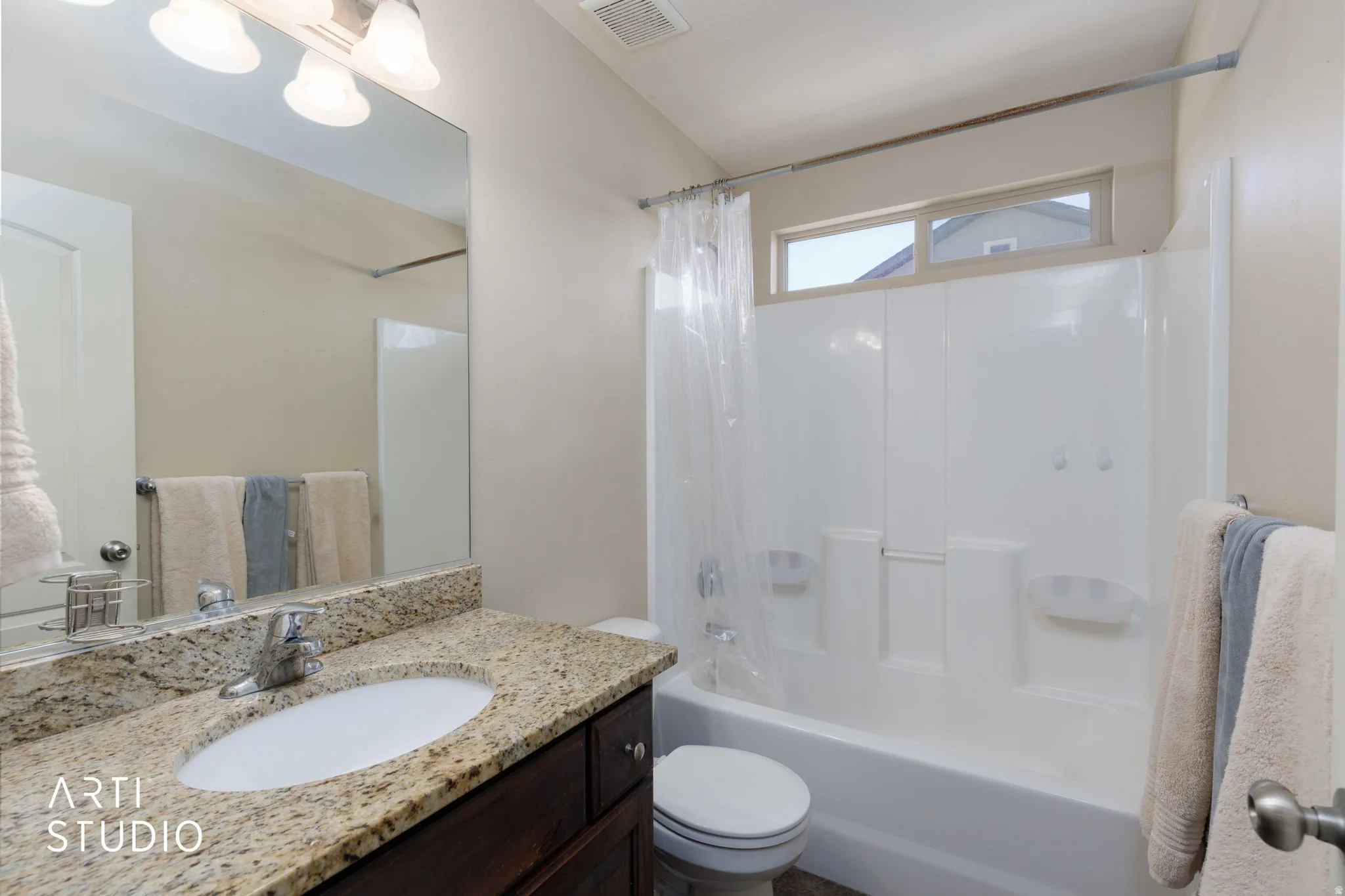 Bathroom featuring a white integrated tub and shower unit with a window above, a dark wood-finish vanity, a stone-finish countertop, and a chrome faucet