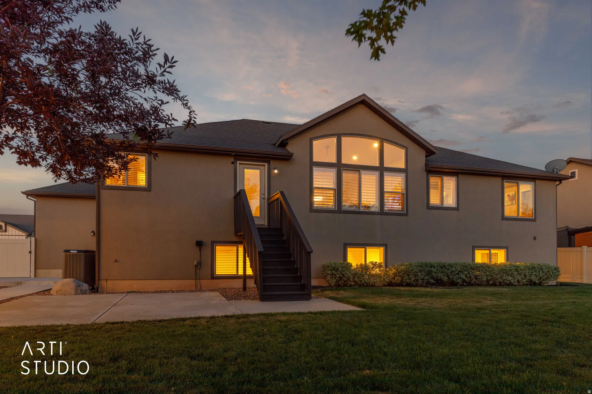 Rear exterior featuring a large arched window, multiple distinct windows, a ground-level patio, and a staircase leading to an upper-level door