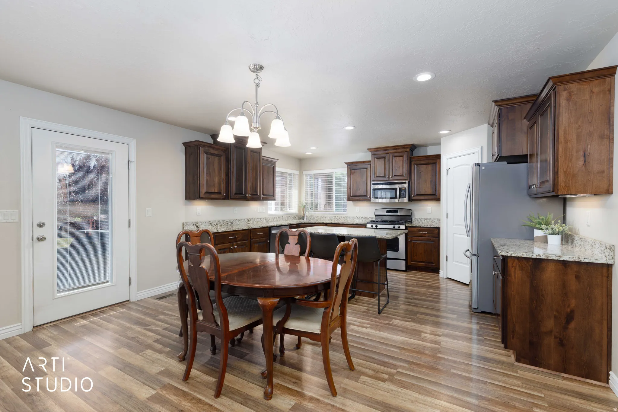 Spacious kitchen and dining area featuring wood-finish flooring, dark wood cabinetry, light granite countertops, and stainless steel appliances