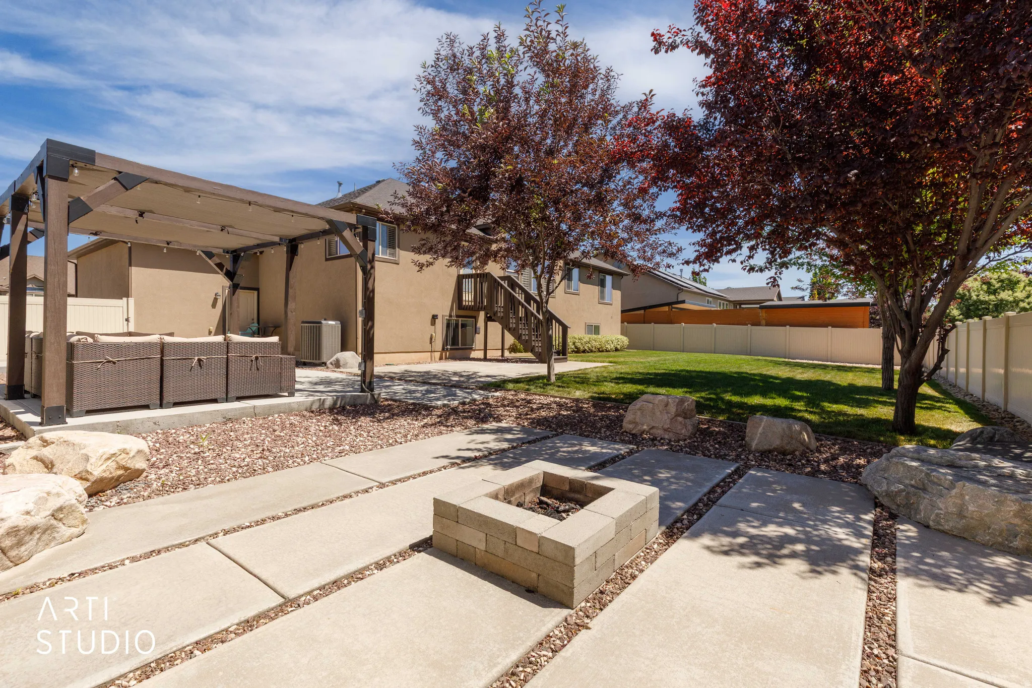 Spacious backyard featuring a pergola-covered patio, a built-in fire pit, concrete paver pathways with decorative gravel, mature trees, and a manicured lawn area