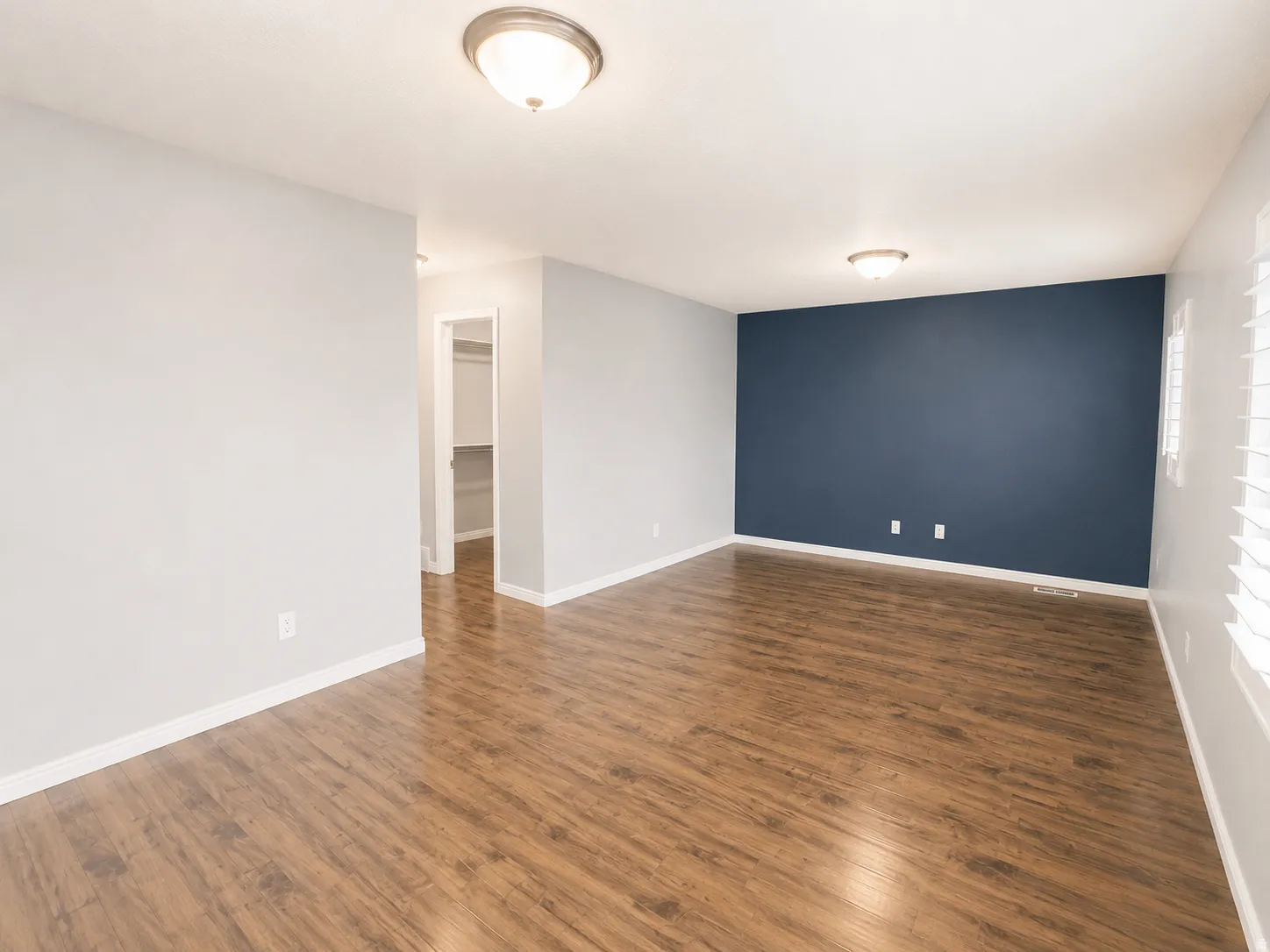 Spacious interior room featuring wood-finish flooring, a contemporary accent wall, light gray painted walls, white trim, and a semi-flush mount ceiling light