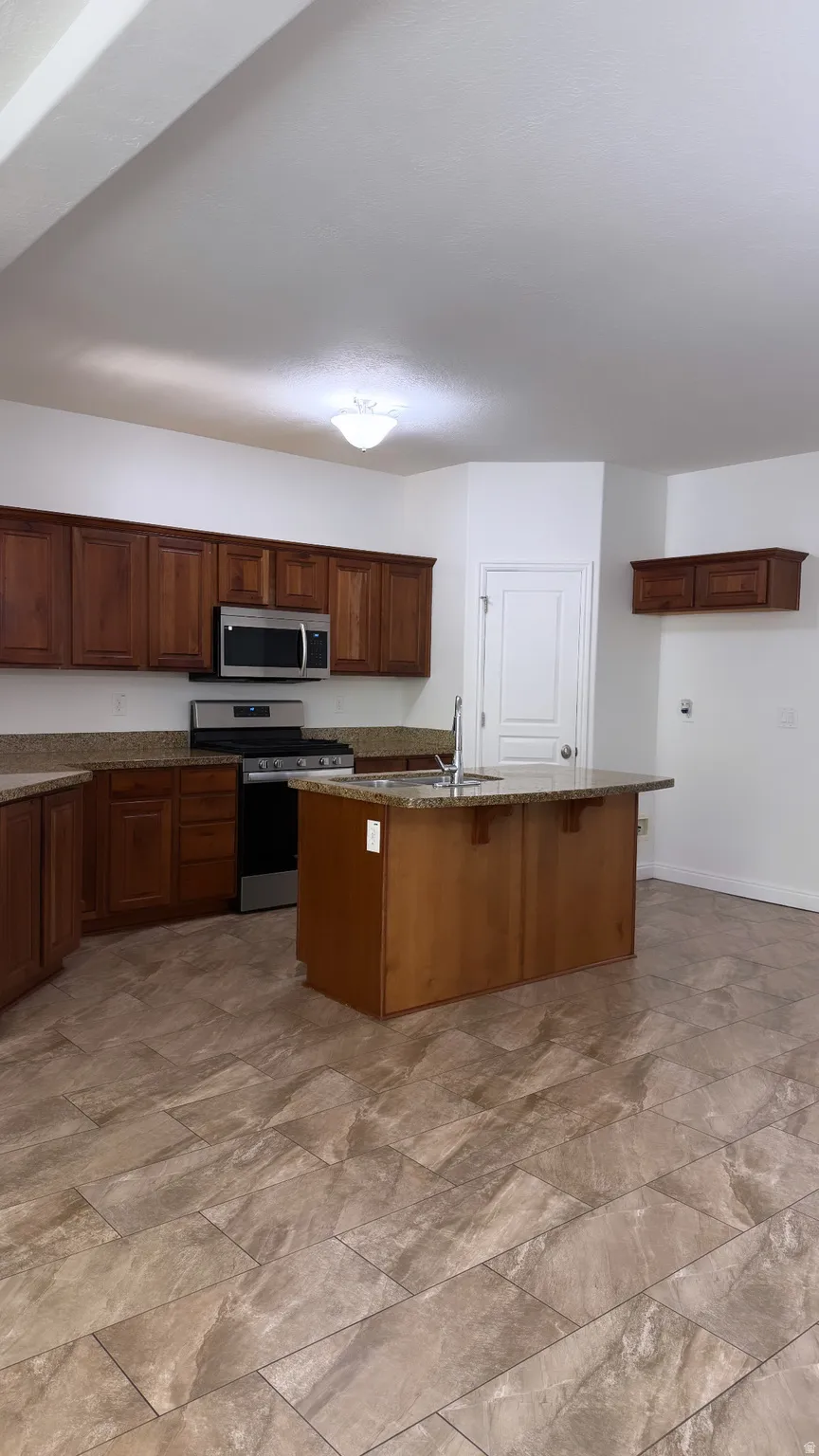 Kitchen featuring wood cabinetry, granite-style countertops, stainless steel appliances, a central island with an integrated sink, and large format tile flooring