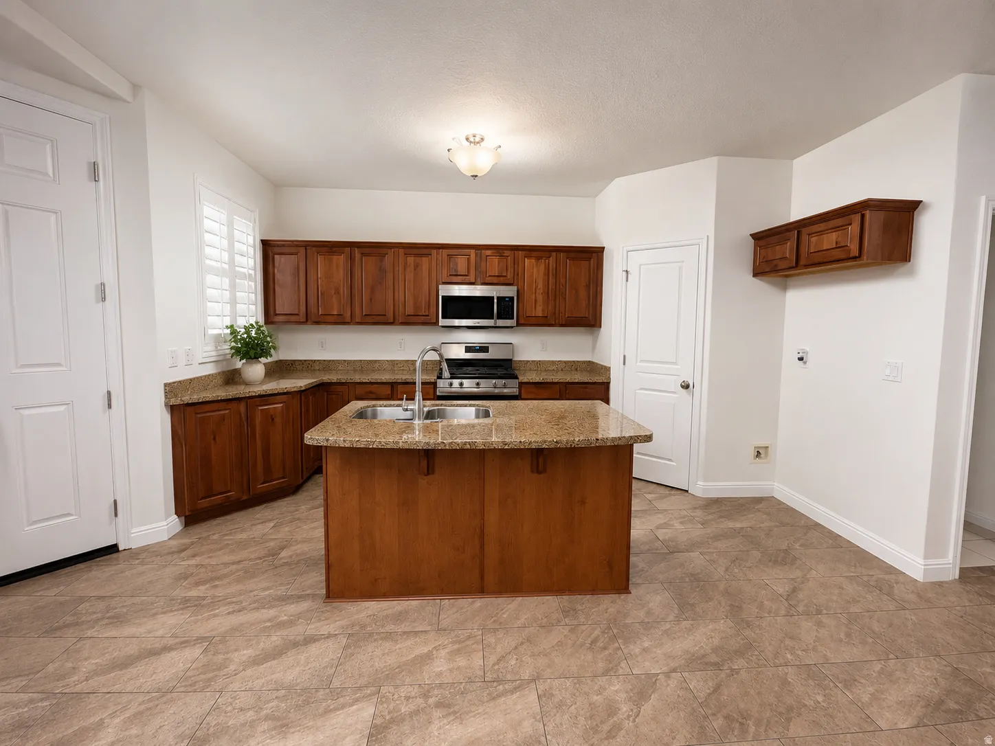 Spacious kitchen featuring a central island with an integrated sink, granite-finish countertops, warm wood cabinetry, stainless steel appliances, and large format tile flooring