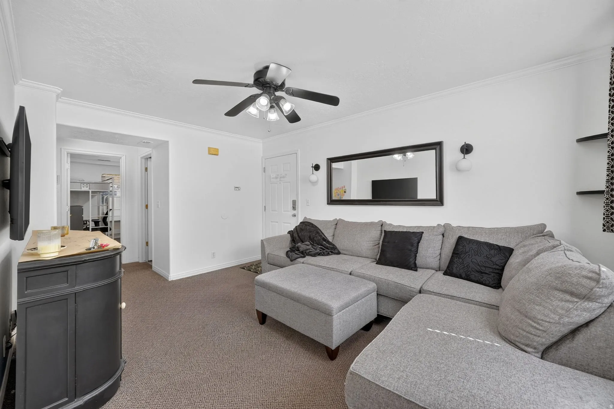 Carpeted living area featuring a ceiling fan with integrated lighting, crown molding, and a white entry door