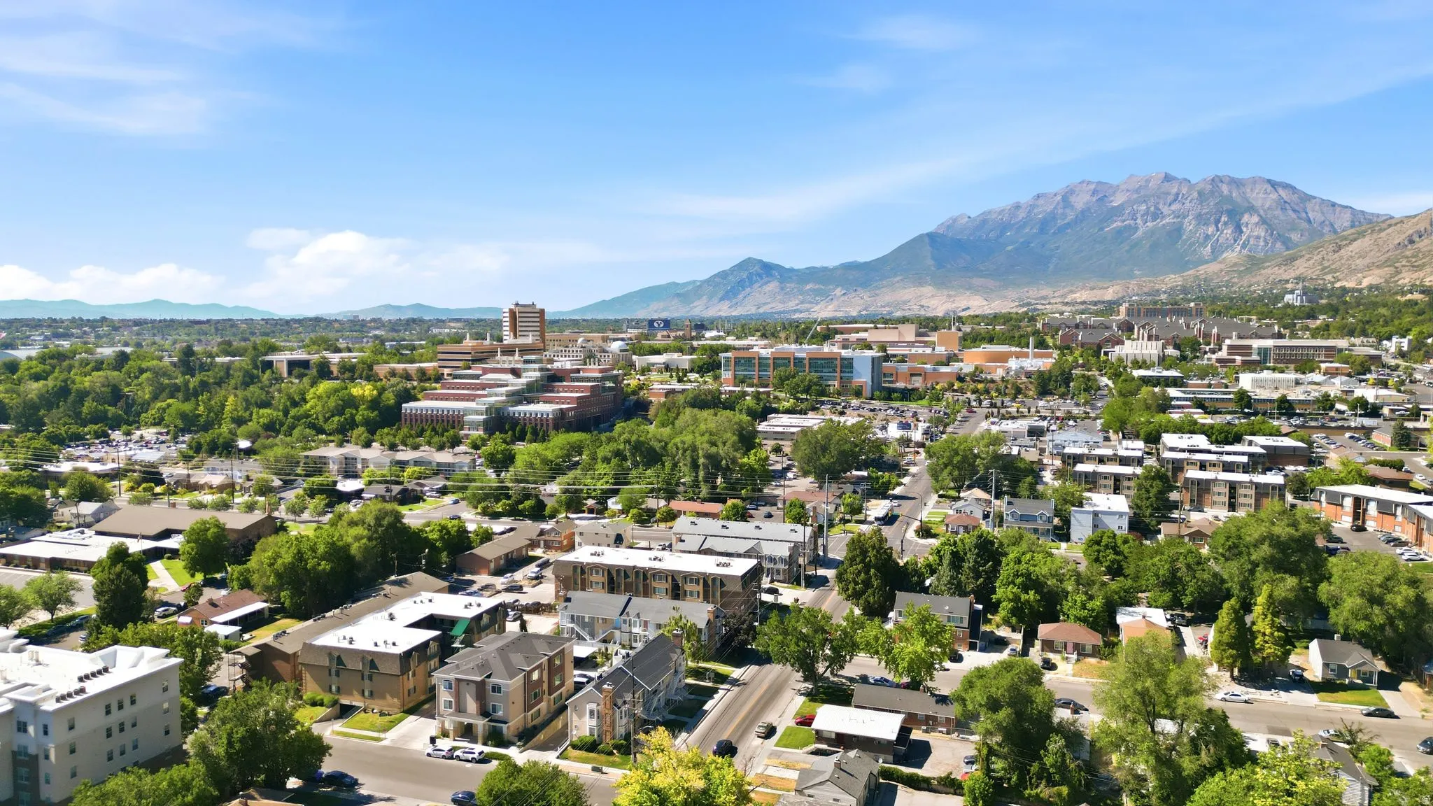 Expansive urban landscape featuring a diverse mix of residential and commercial buildings, lush green trees, and distant mountains under a clear blue sky