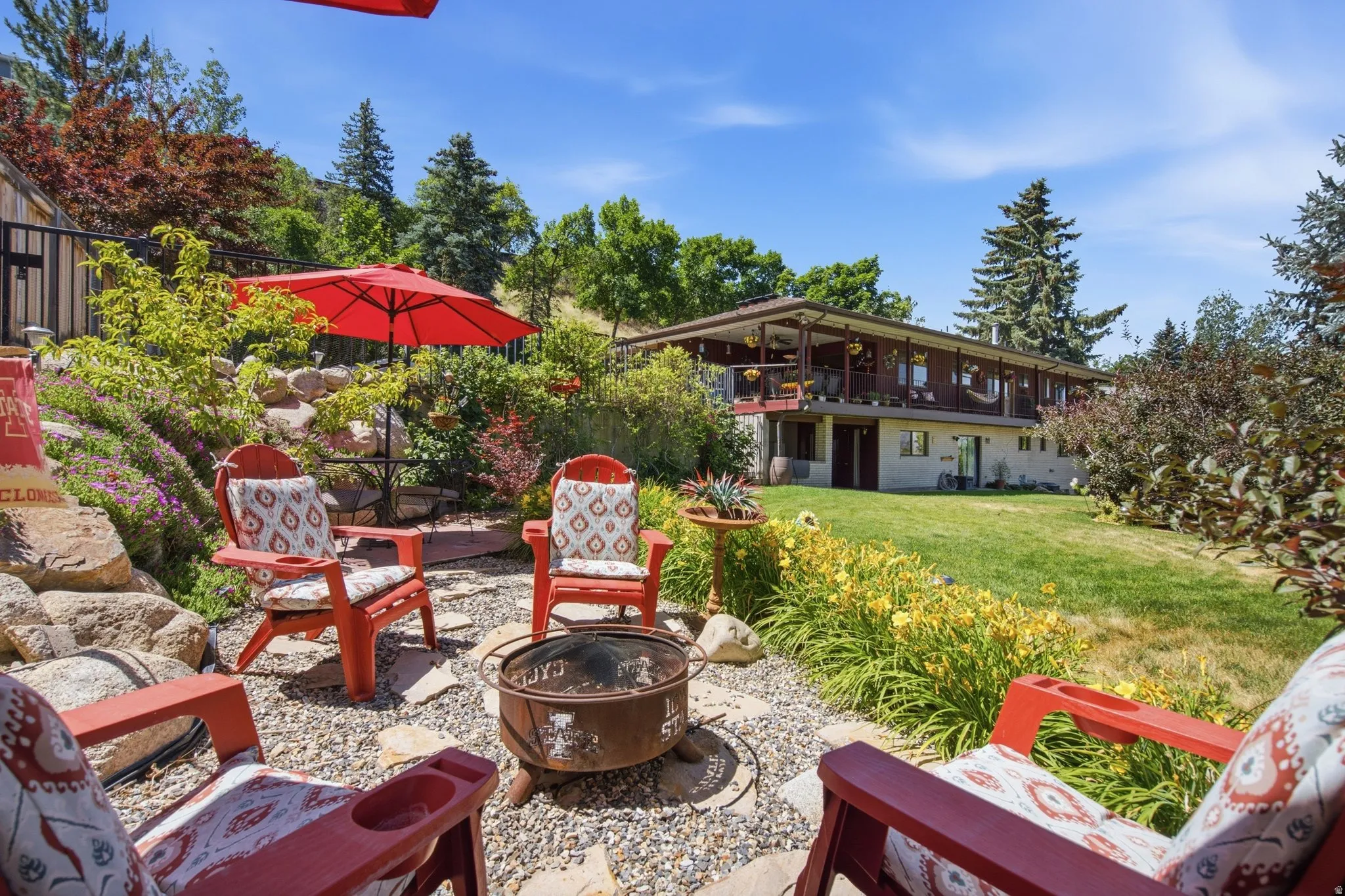 Terraced backyard featuring a gravel patio with a built-in fire pit, stone landscaping, mature trees, and perennial plantings