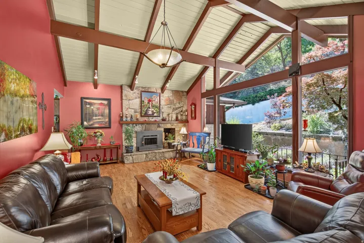 Vaulted ceiling with exposed wood beams, expansive floor-to-ceiling windows, natural stone fireplace with wood-burning insert, wood-finish flooring, and a contemporary chandelier