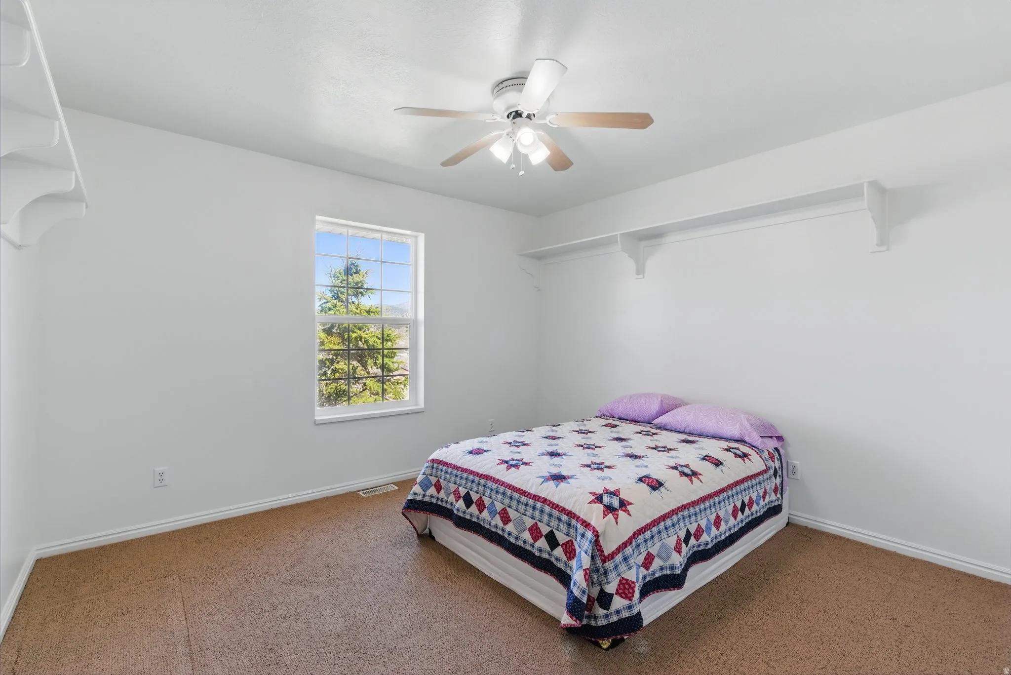Carpeted room featuring a ceiling fan with light fixture, a window with exterior foliage visible, and built-in shelving