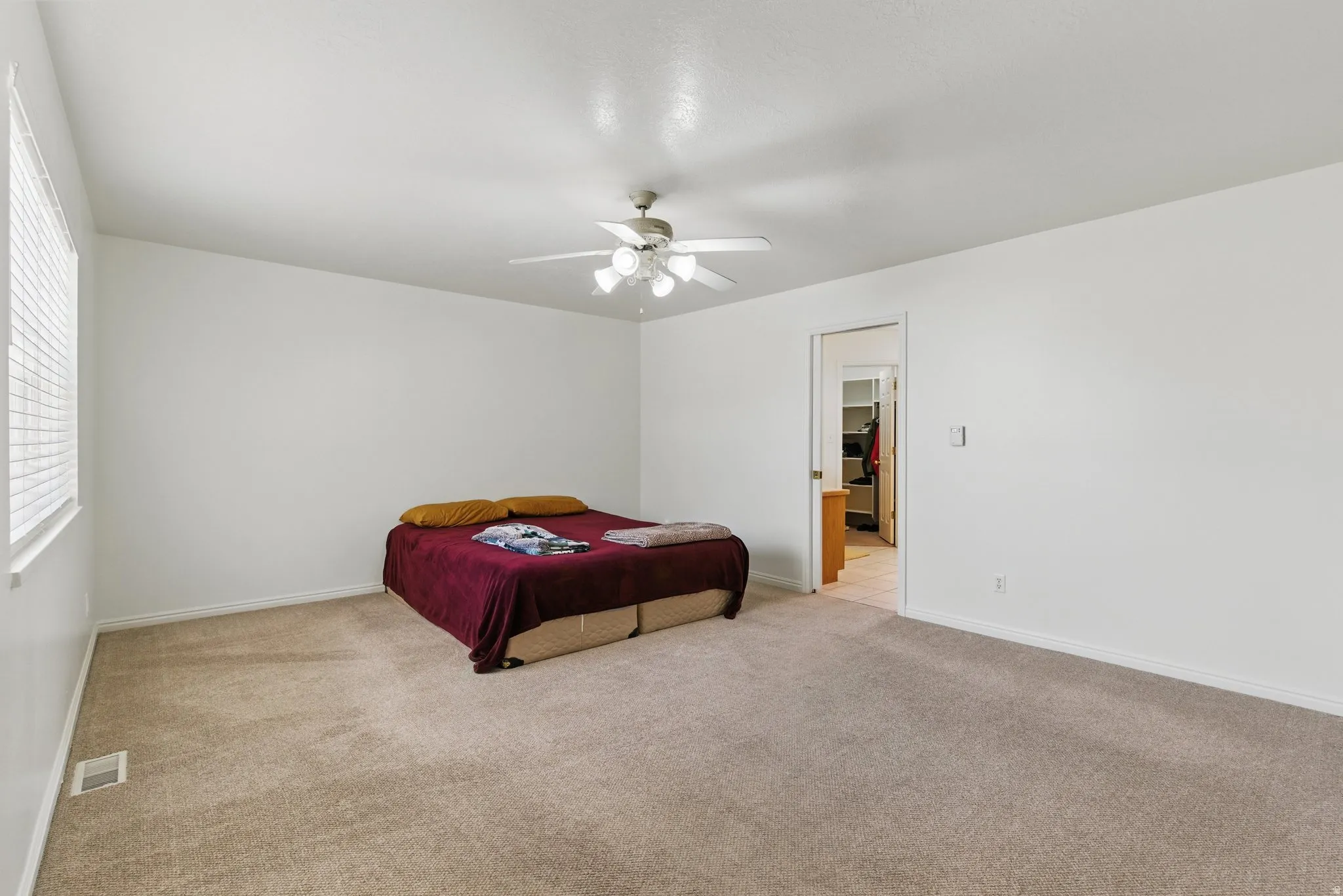Spacious interior room featuring light beige carpeting, white walls, a ceiling fan with integrated lighting, and natural light from a window with blinds