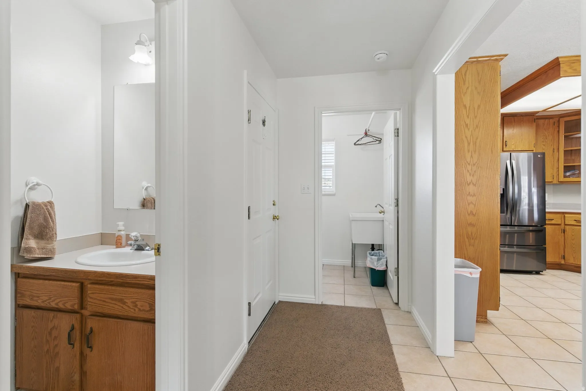 Hallway featuring tile flooring, leading to a utility room with a utility sink and a partial bathroom with a wood-finish vanity and white sink
