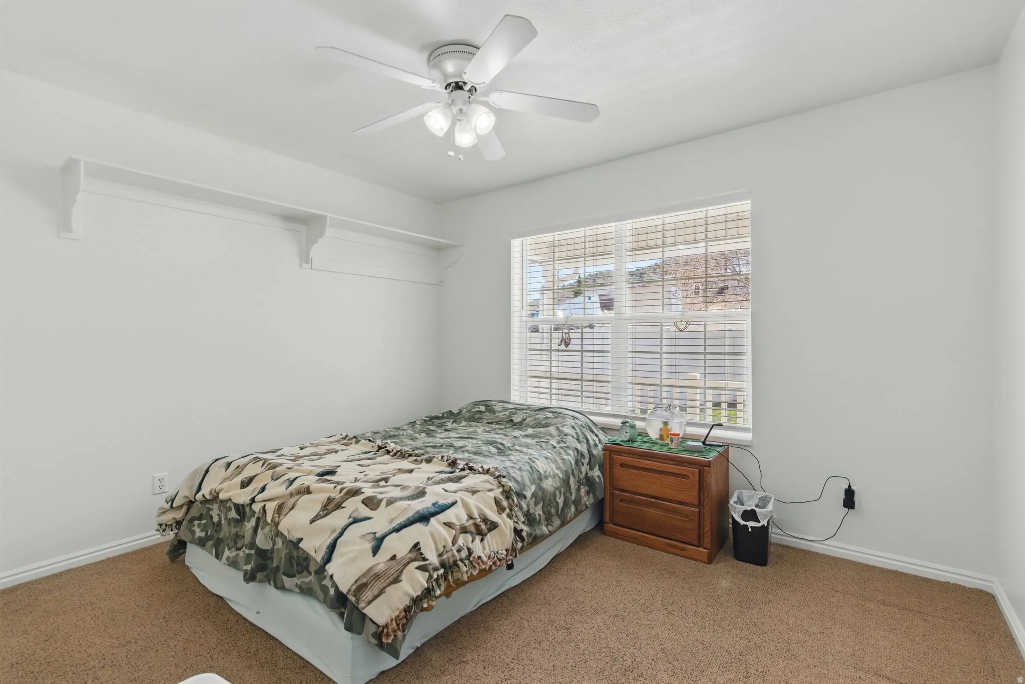 Carpeted room featuring a white ceiling fan with integrated lighting, a large window with horizontal blinds, and a built-in shelf system
