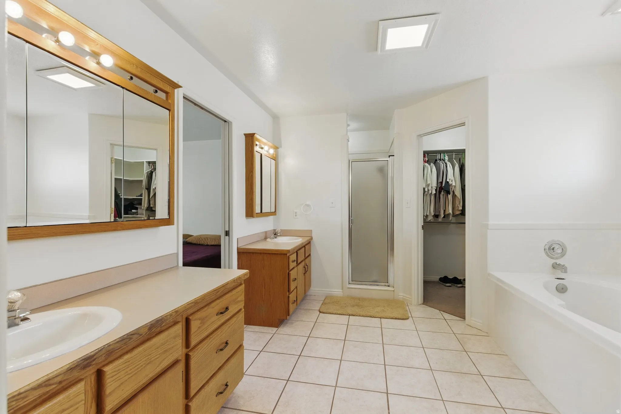 Bathroom featuring dual wood-finish vanities with integrated sinks, a built-in tub, a glass-enclosed shower, and neutral tile flooring