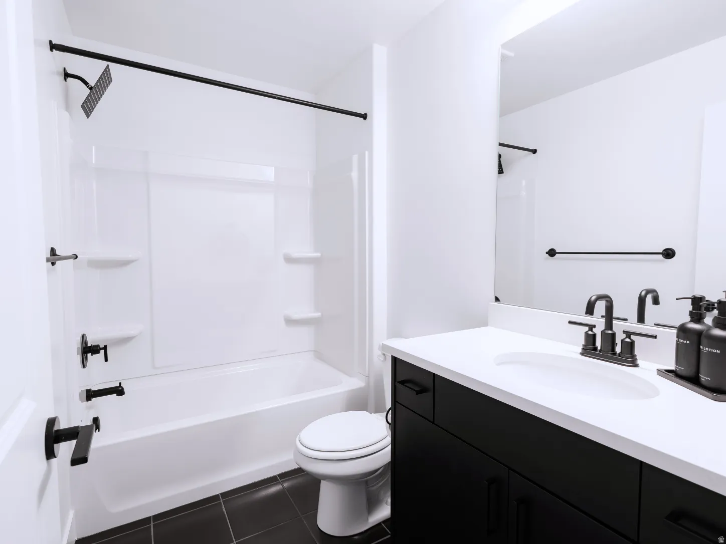 Bathroom featuring a white tub/shower combination, black shower fixtures, a white toilet, a dark vanity with a white countertop and integrated sink, and a framed mirror
