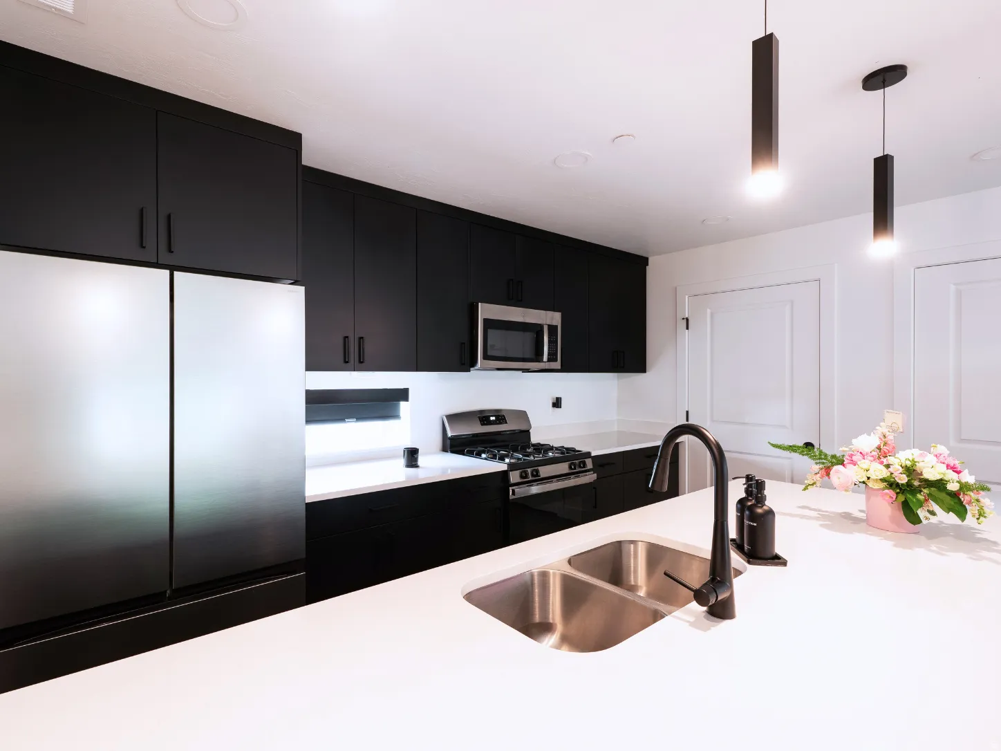 Contemporary kitchen featuring stainless steel appliances, black flat-panel cabinetry, white countertops, a dual basin sink with a matte black faucet, and pendant lighting