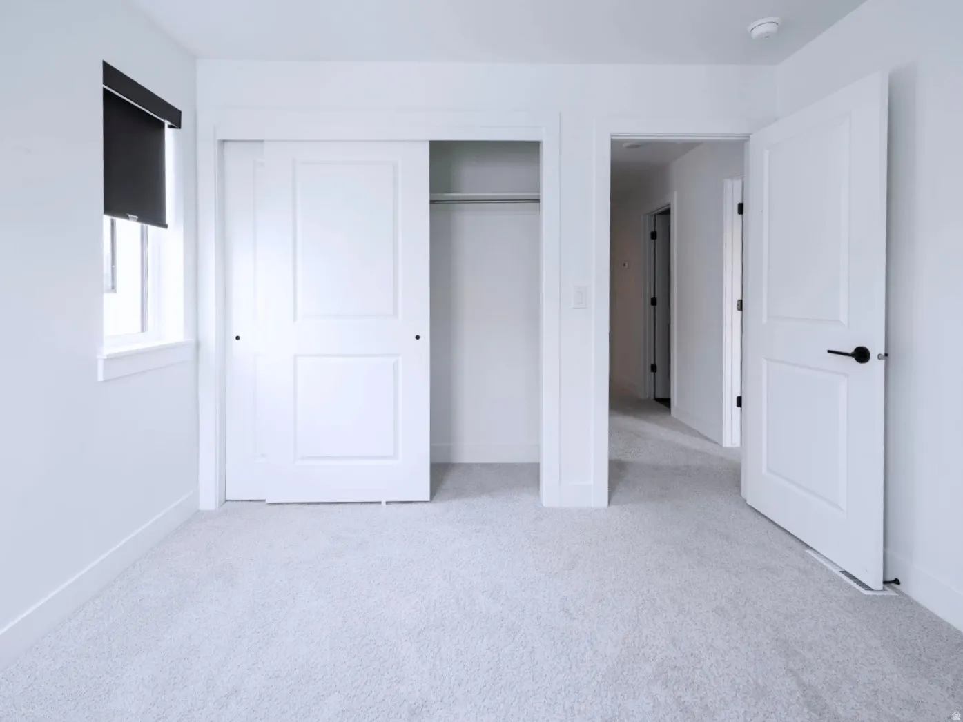 Bright interior room featuring light-colored carpeting, white walls, and a window with a dark roller blind