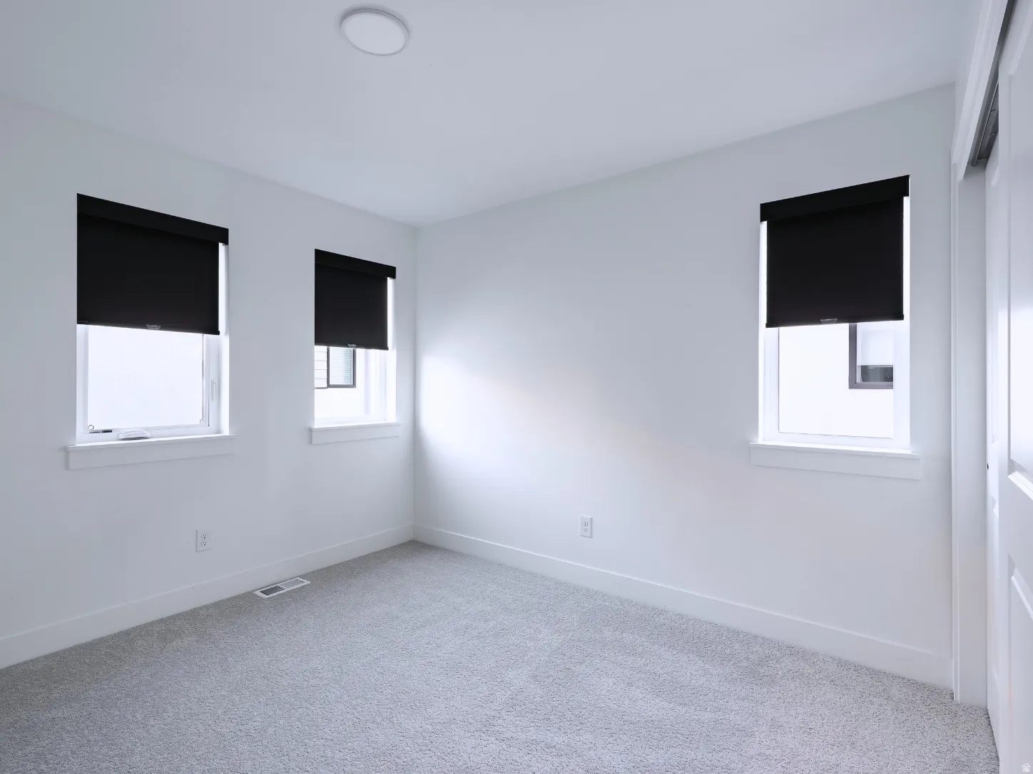 Bright room featuring three windows with roller shades, light grey carpeting, and white walls
