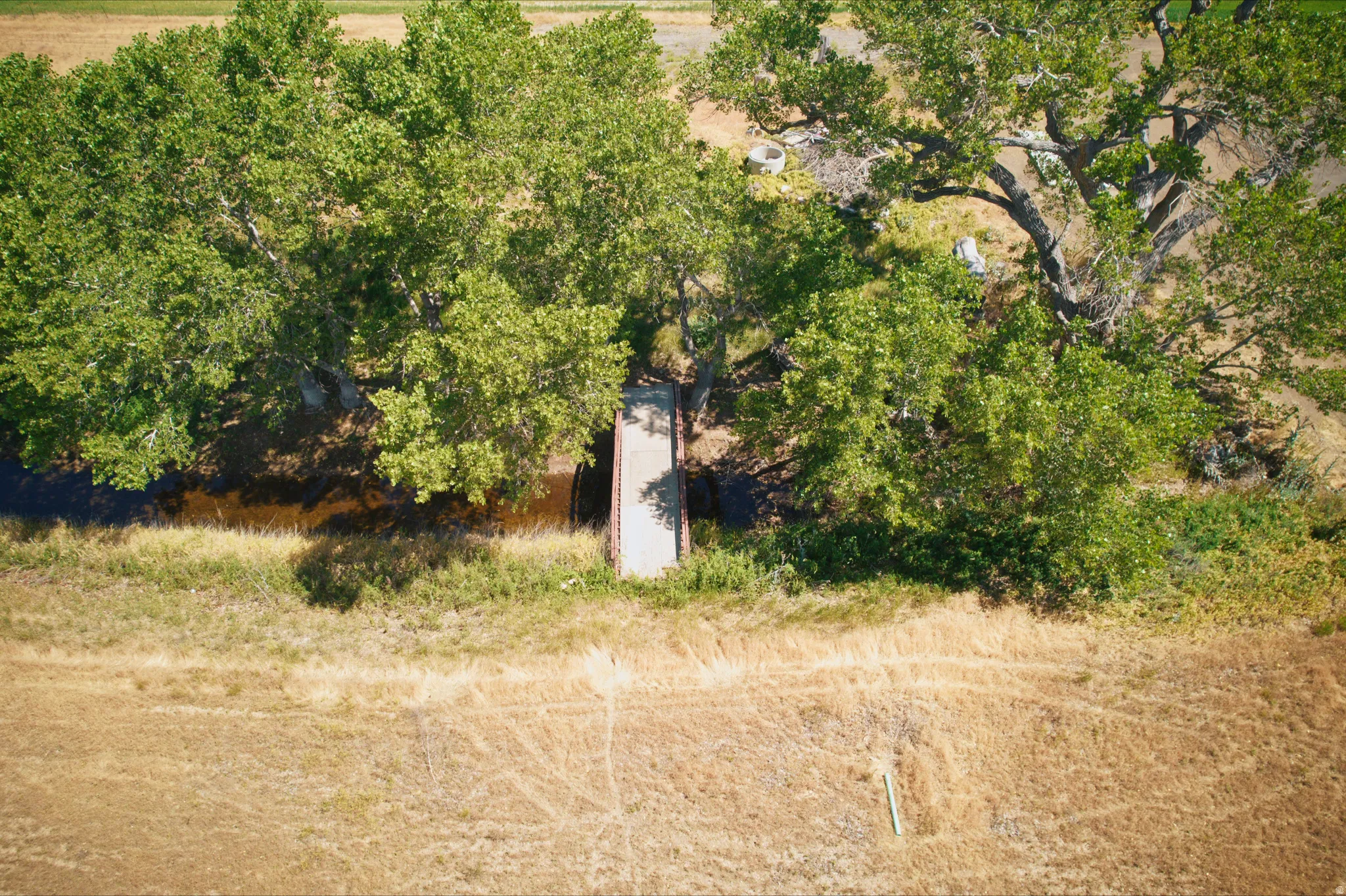 Aerial view showcasing a natural water feature with a bridge crossing, bordered by mature trees and expansive dry grass areas
