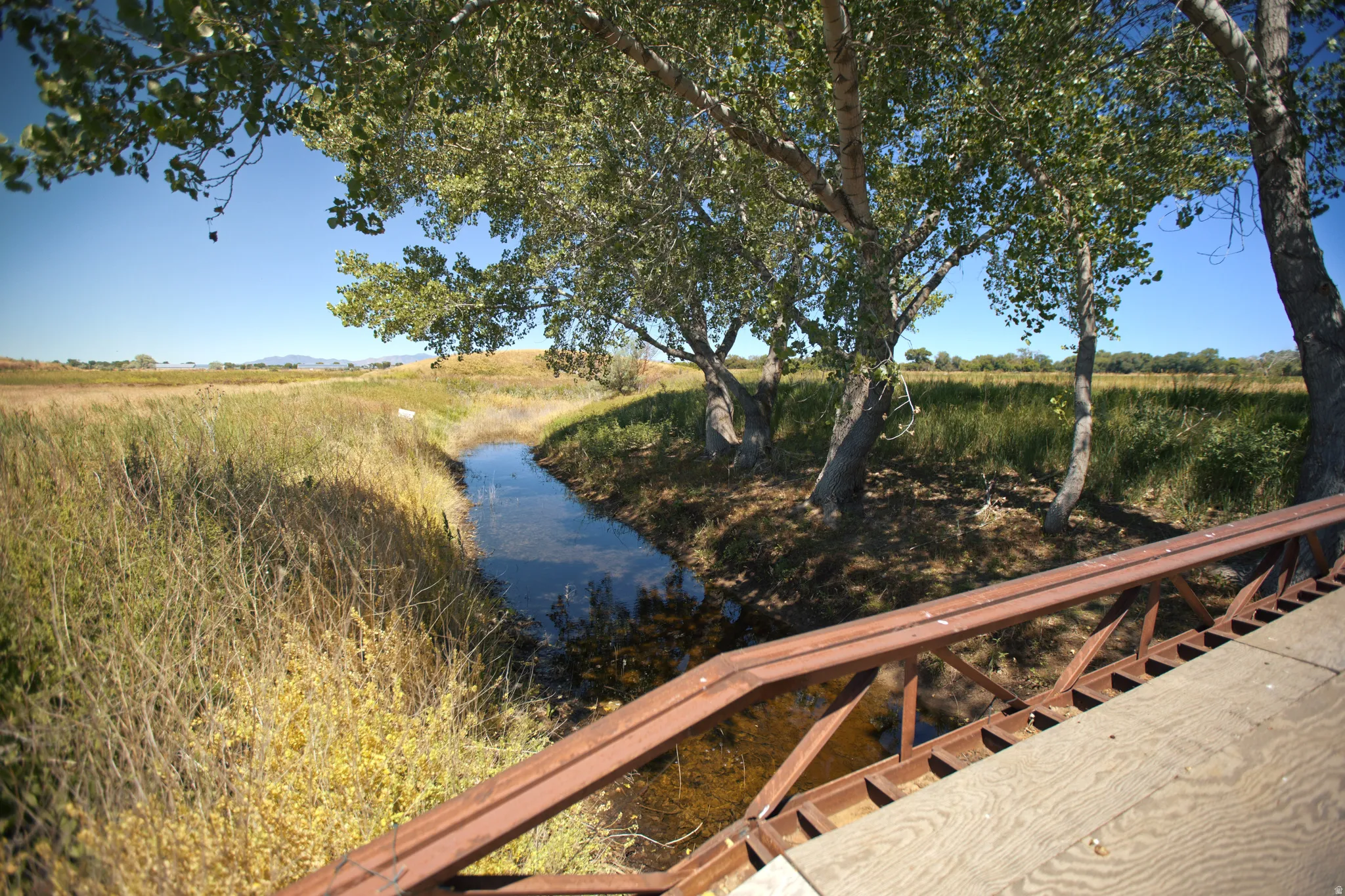 Expansive natural landscape featuring a waterway, mature trees, and a rustic bridge with a metal frame and wood deck