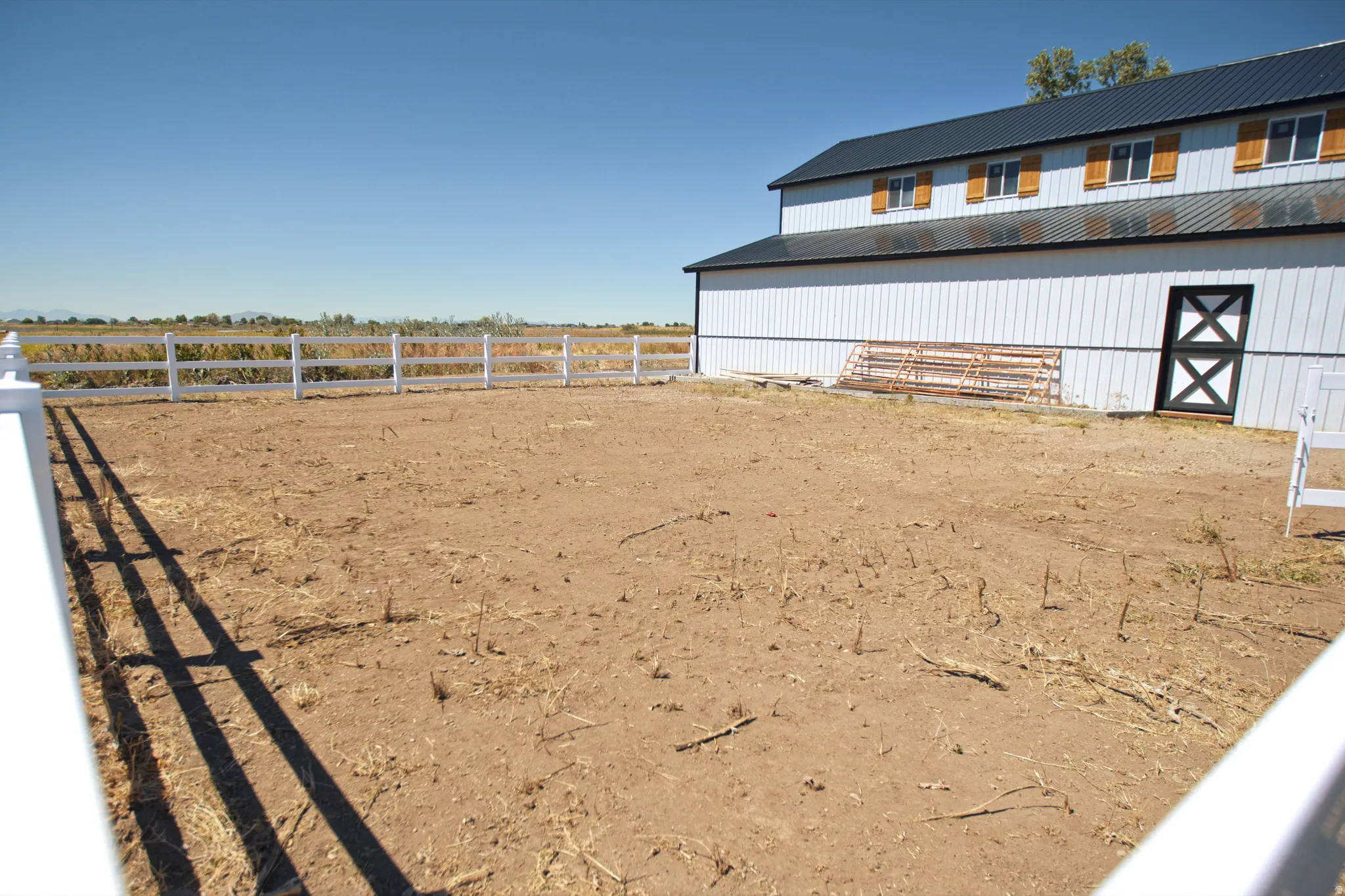 Expansive outdoor area with a white two-rail fence, a metal-roofed barn-style structure featuring white vertical siding, multiple windows with natural wood shutters, and a black and white crossbuck door
