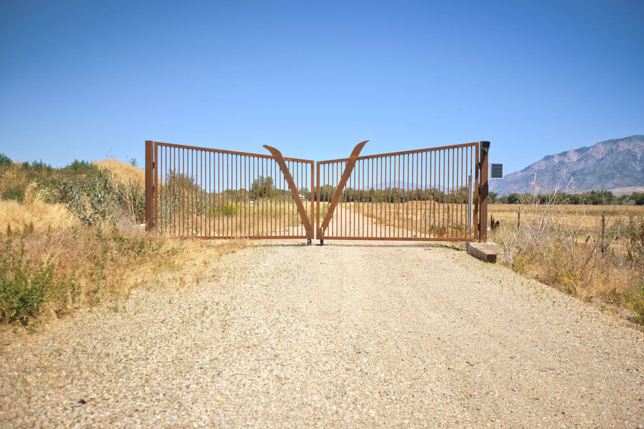 Expansive double-gated entry featuring a gravel drive, sturdy metal gates, and a natural landscape backdrop