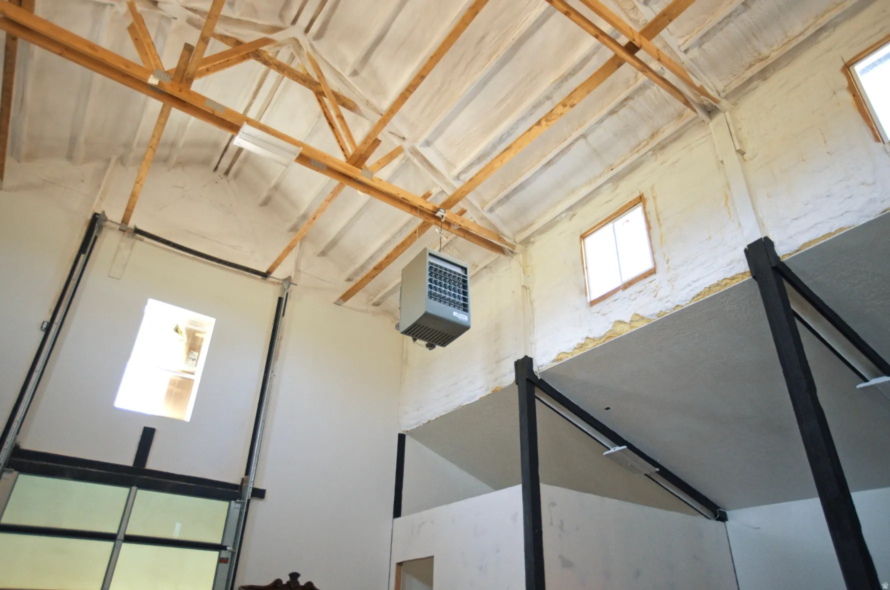 High vaulted ceiling with exposed wood trusses, spray foam insulation, multiple windows, and a large overhead door with glass panels