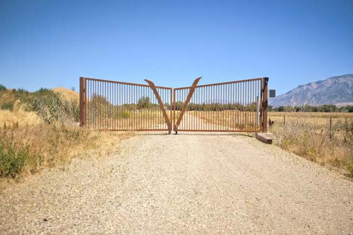 Expansive double-gated entry featuring a gravel drive, sturdy metal gates, and a natural landscape backdrop