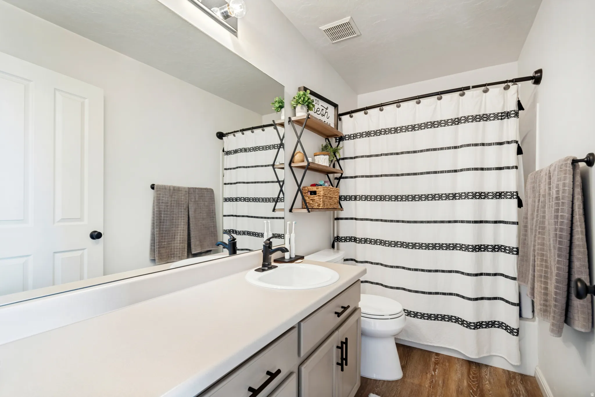 Bathroom featuring wood-finish flooring, a white vanity with a single basin sink and dark-finish faucet, a wall-mounted mirror, and a white tub/shower combination