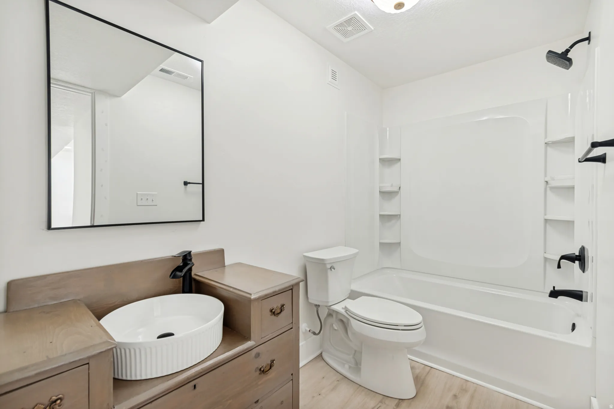 Modern bathroom featuring a wood-finish vanity with an oval vessel sink, matte black fixtures, a rectangular framed mirror, a built-in bathtub with shower, and light wood-finish flooring