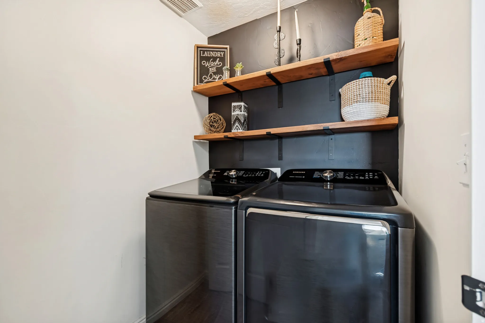 Dedicated laundry space featuring a dark accent wall, two wood-finish floating shelves, and wood-finish flooring