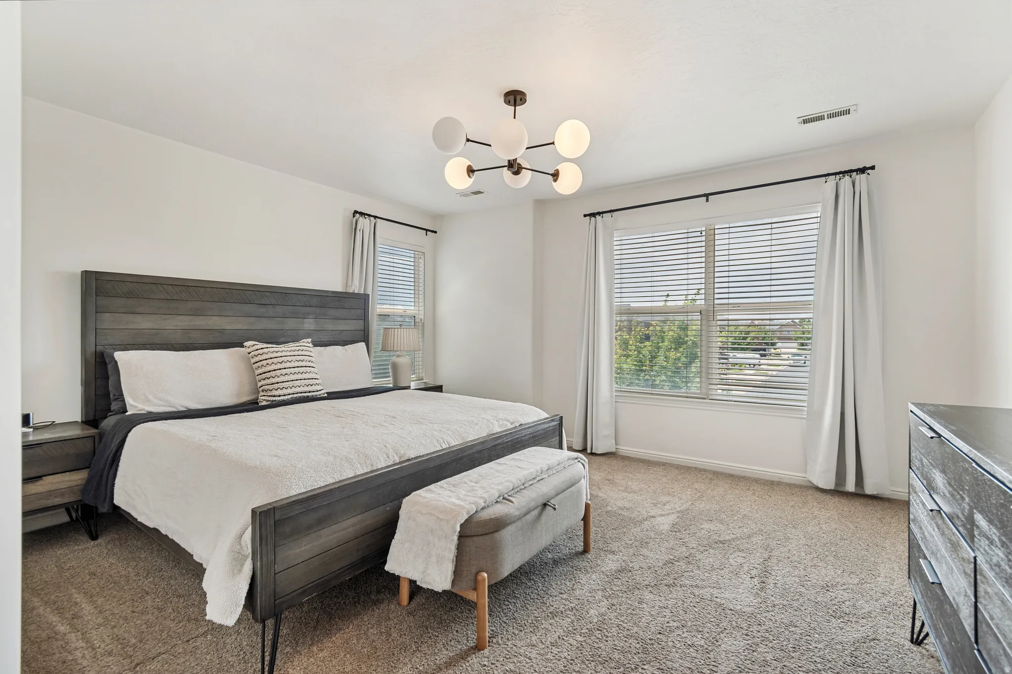 Spacious room featuring a modern spherical chandelier, soft carpeting, and dual windows with blinds and drapes
