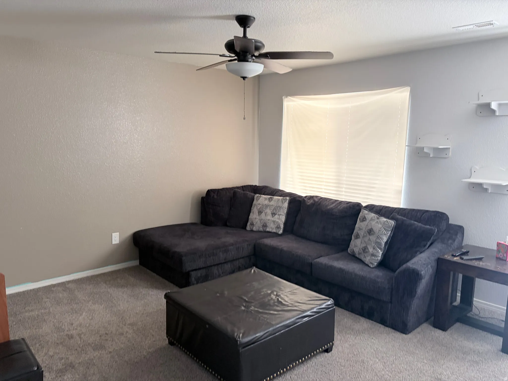 Carpeted living space featuring a ceiling fan with integrated lighting, a window with horizontal blinds, and wall-mounted shelving
