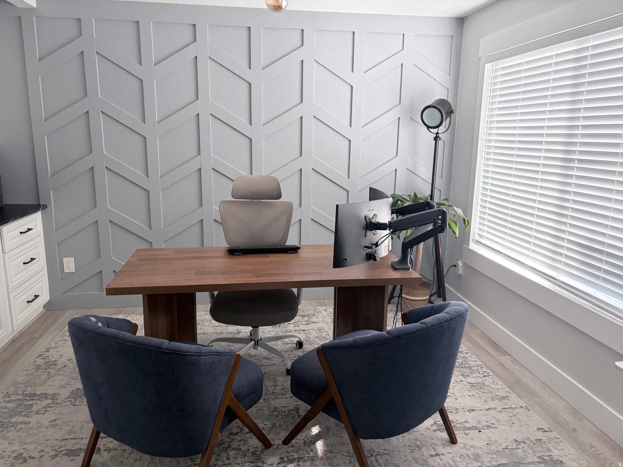 Accent wall featuring geometric paneling, wood-finish flooring, a white trim window with blinds, and built-in white cabinetry with black hardware