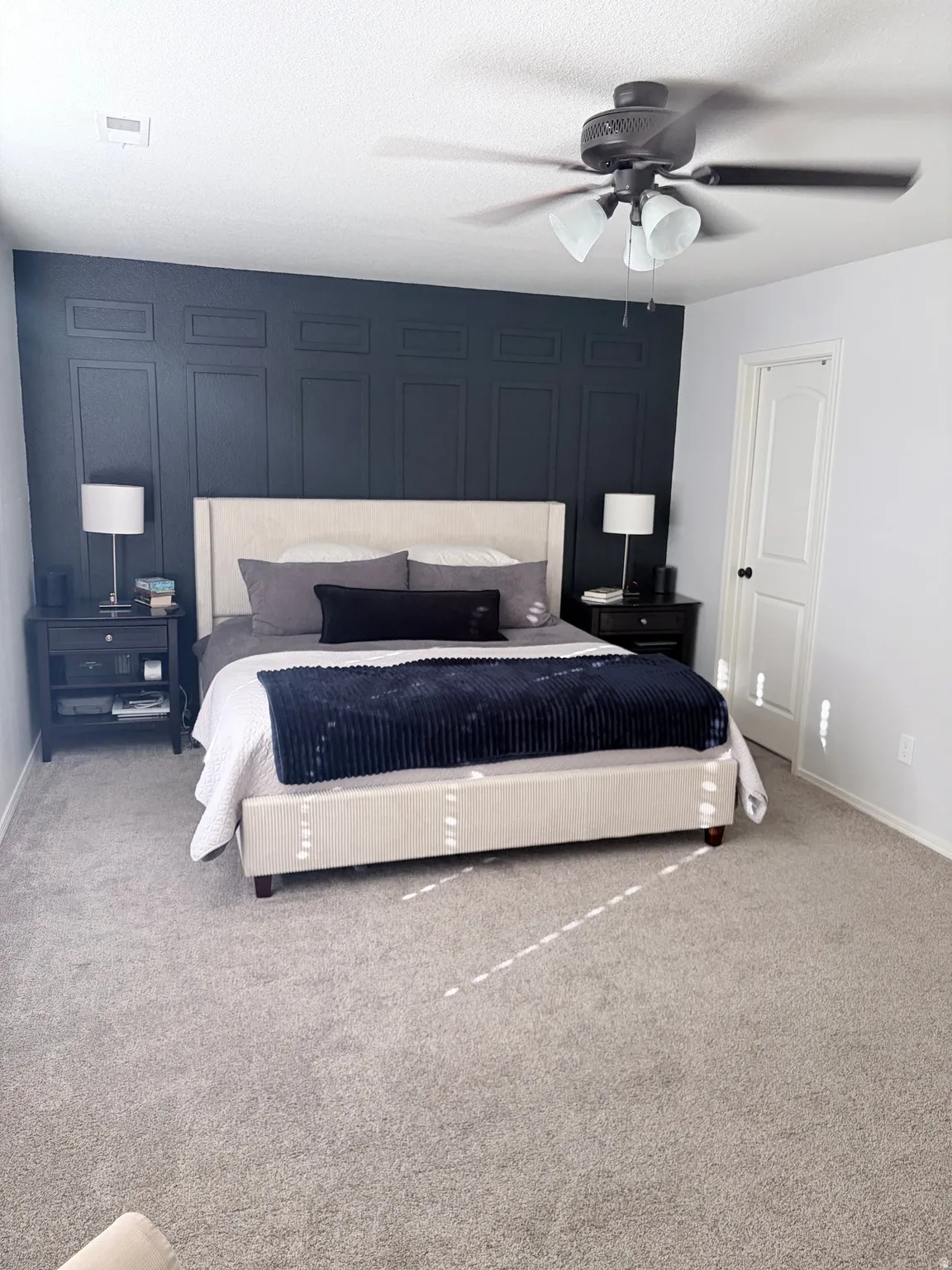 Spacious bedroom featuring dark paneled accent wall, light grey carpeting, and a ceiling fan with integrated lighting