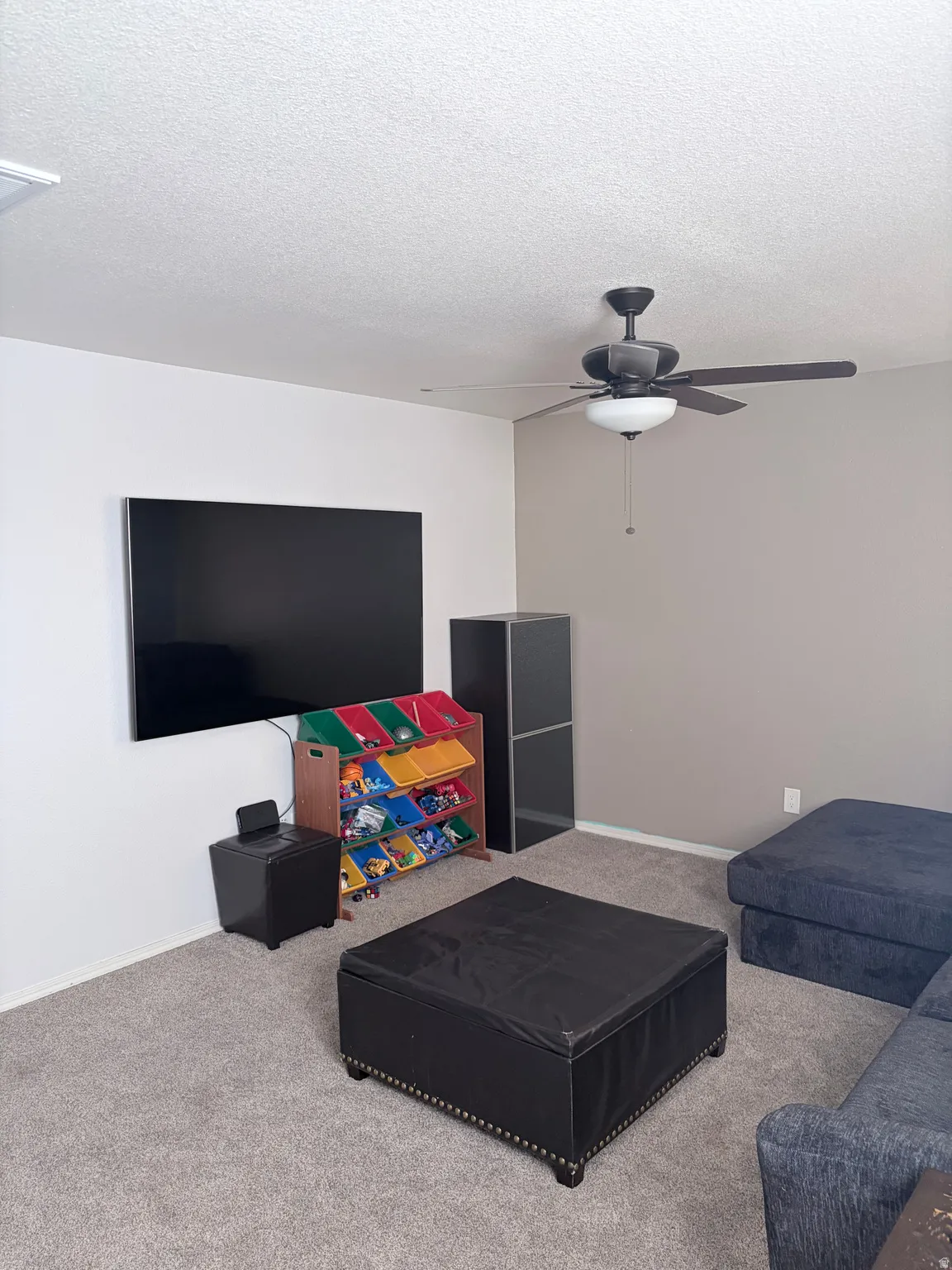 Carpeted room featuring a ceiling fan with light, a wall-mounted display, and neutral-toned walls
