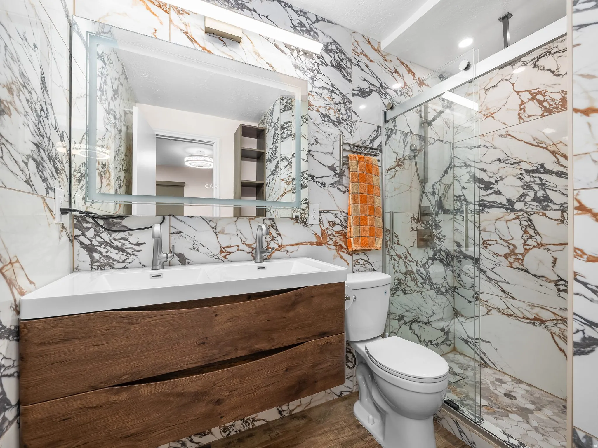 Full-wall marble-finish tiling, a floating wood-finish vanity with an integrated double sink, a large back-lit mirror, a glass-enclosed shower, and hexagonal mosaic shower flooring