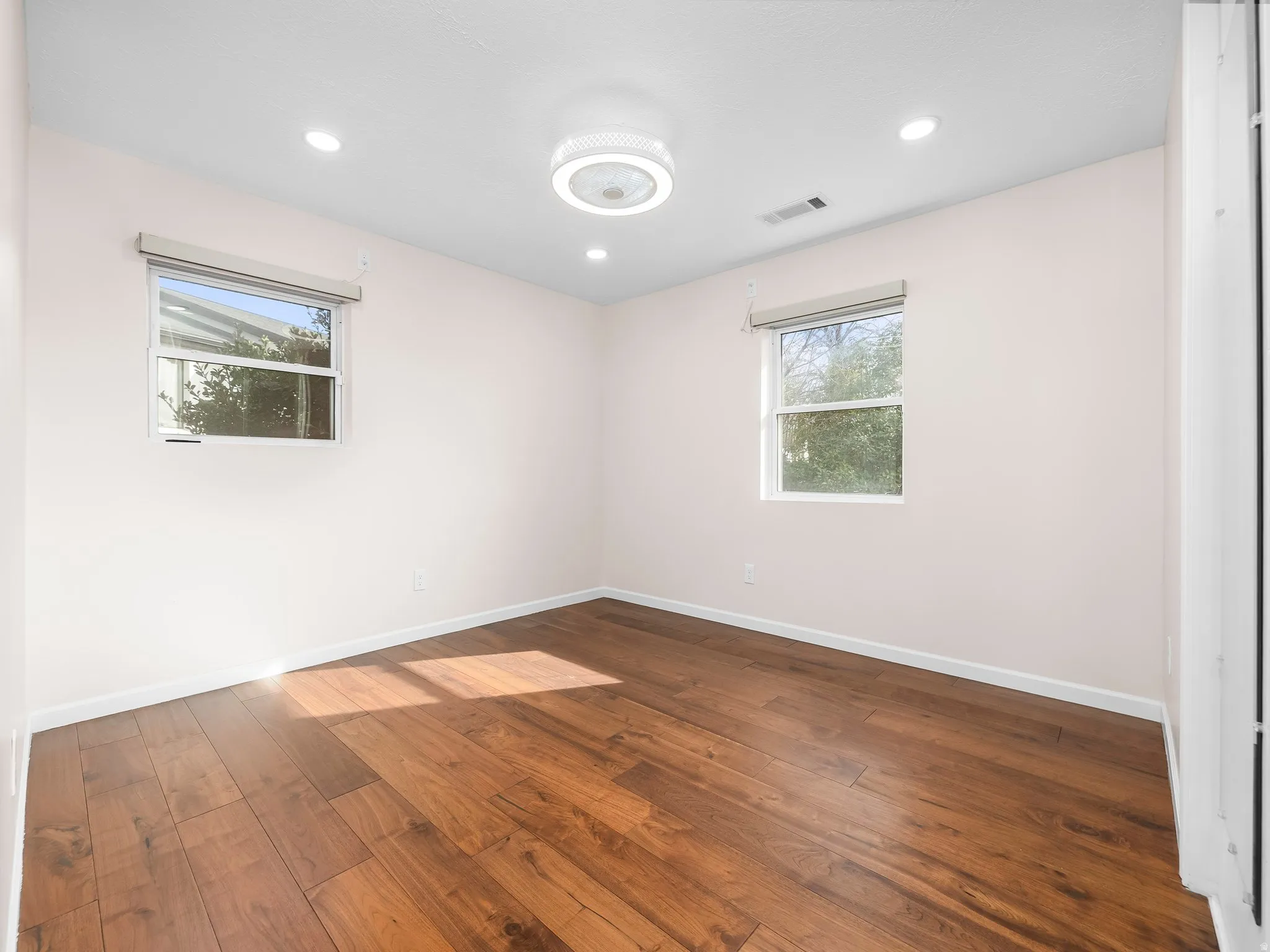 Bright room featuring wood-finish flooring, two windows with roller blinds, recessed lighting, and a modern ceiling fan