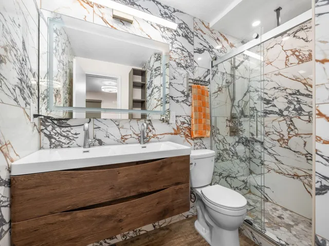 Full-wall marble-finish tiling, a floating wood-finish vanity with an integrated double sink, a large back-lit mirror, a glass-enclosed shower, and hexagonal mosaic shower flooring