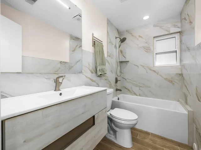 Bathroom featuring a wood-finish floating vanity with an integrated sink, brushed nickel faucet, full-width mirror, white bathtub with a shower system, and ceramic tile walls extending to the ceiling