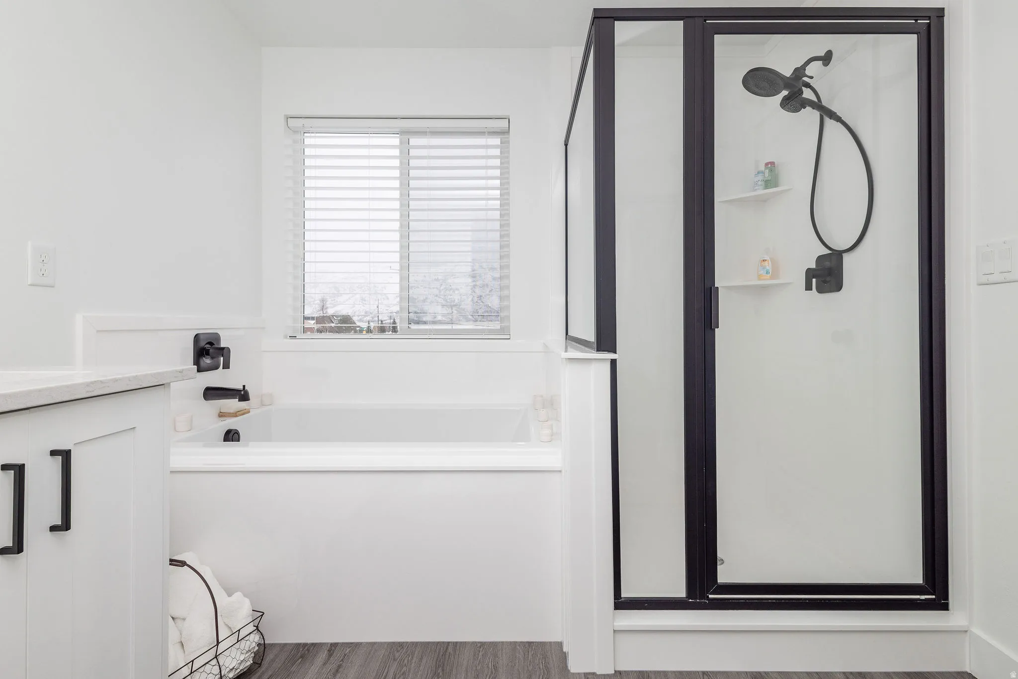 Contemporary bathroom featuring a walk-in shower with matte black fixtures, a built-in tub with matte black fixtures, white cabinetry with black hardware, a light-toned countertop, and wood-finish flooring