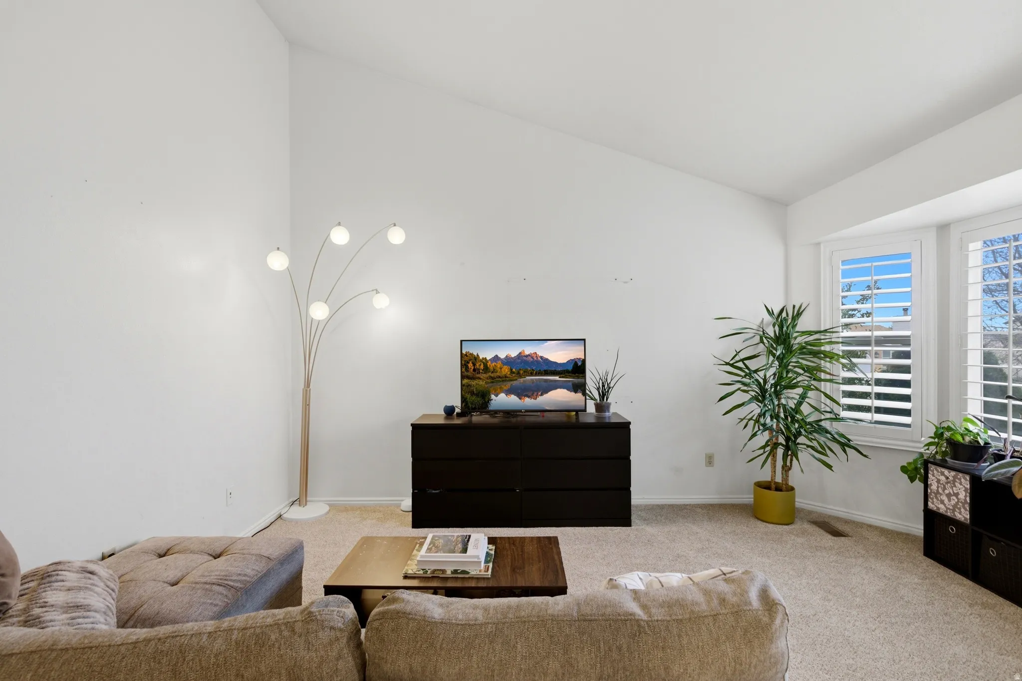 Living area featuring a vaulted ceiling, light neutral carpeting, and a bay window with plantation shutters