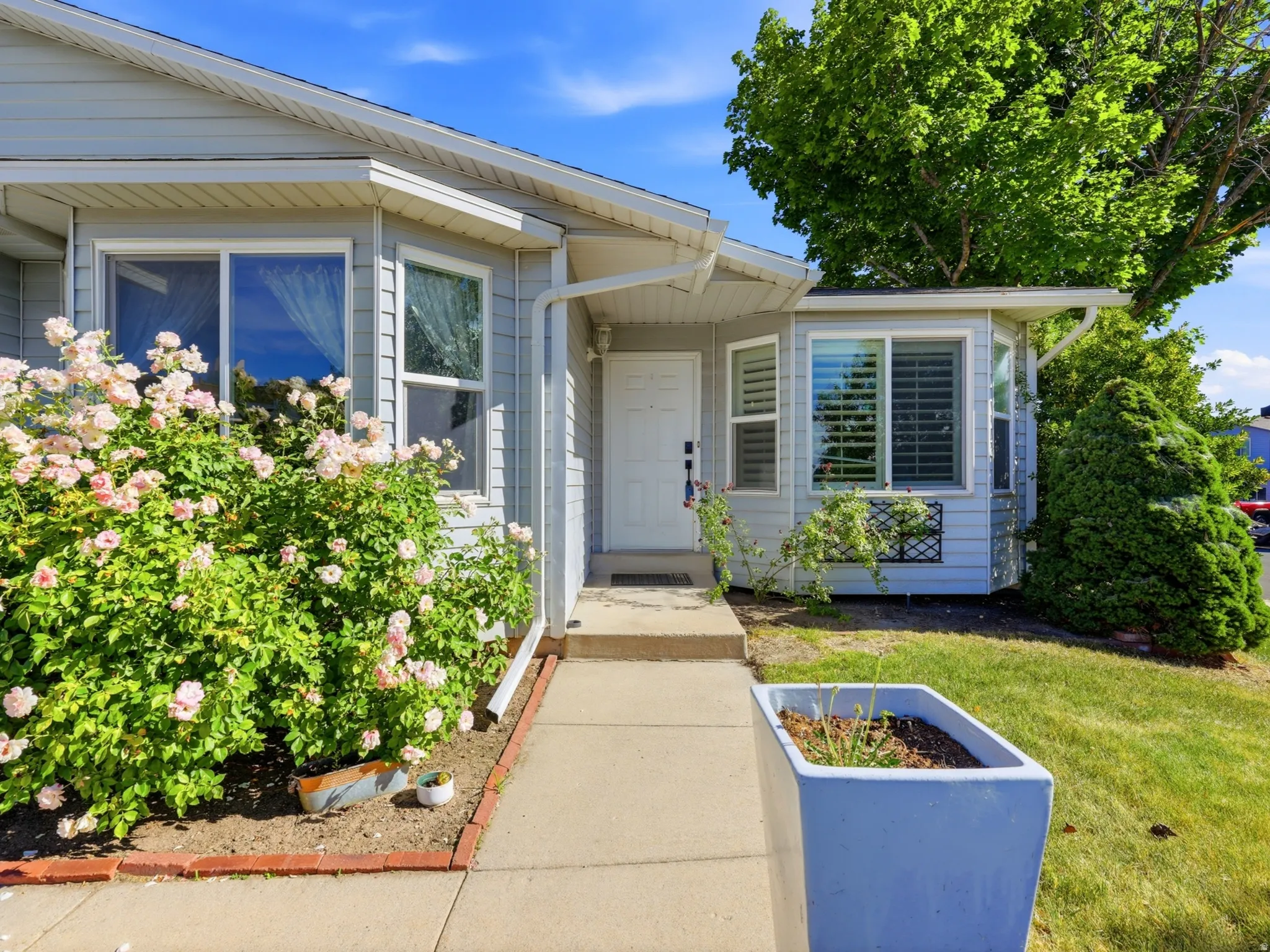 Light grey siding exterior featuring a covered entryway, a prominent bay window with white plantation shutters, and mature landscaping with flowering bushes
