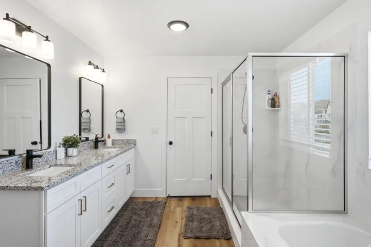 Spacious bathroom featuring a double vanity with granite countertops, wood-finish flooring, a glass-enclosed shower, and a built-in tub