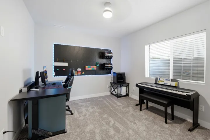 Neutral wall color, light gray carpet flooring, large horizontal window with white blinds, recessed lighting fixture, and a built-in wall-mounted pegboard system with perimeter lighting