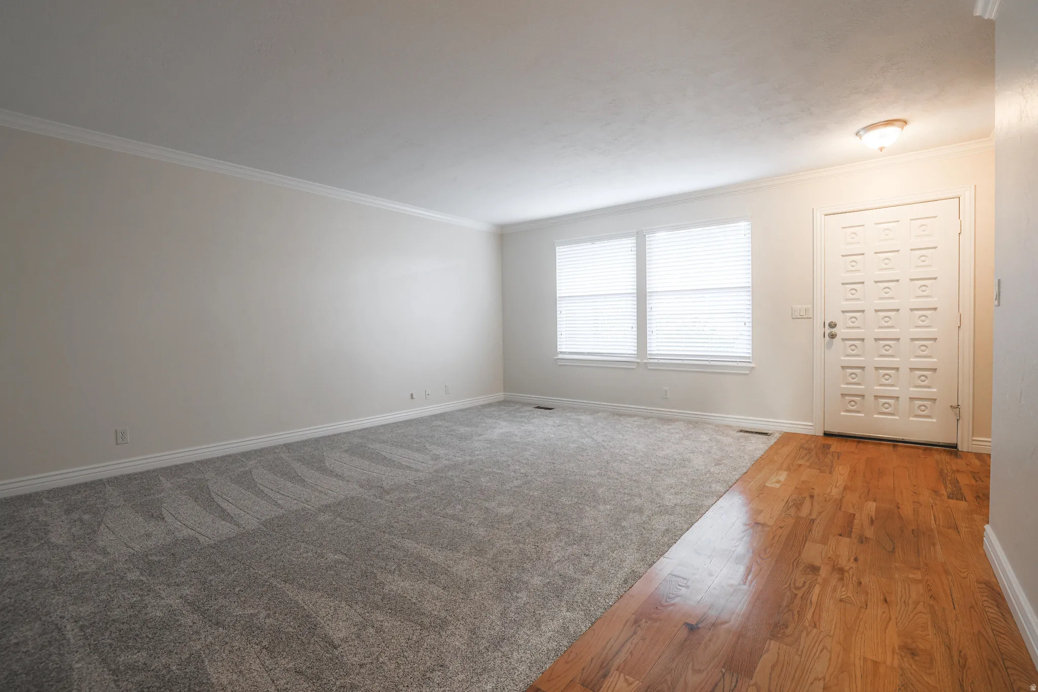Spacious interior featuring a decorative front entry door, two large windows with blinds, a combination of hardwood and carpet flooring, and crown molding accents