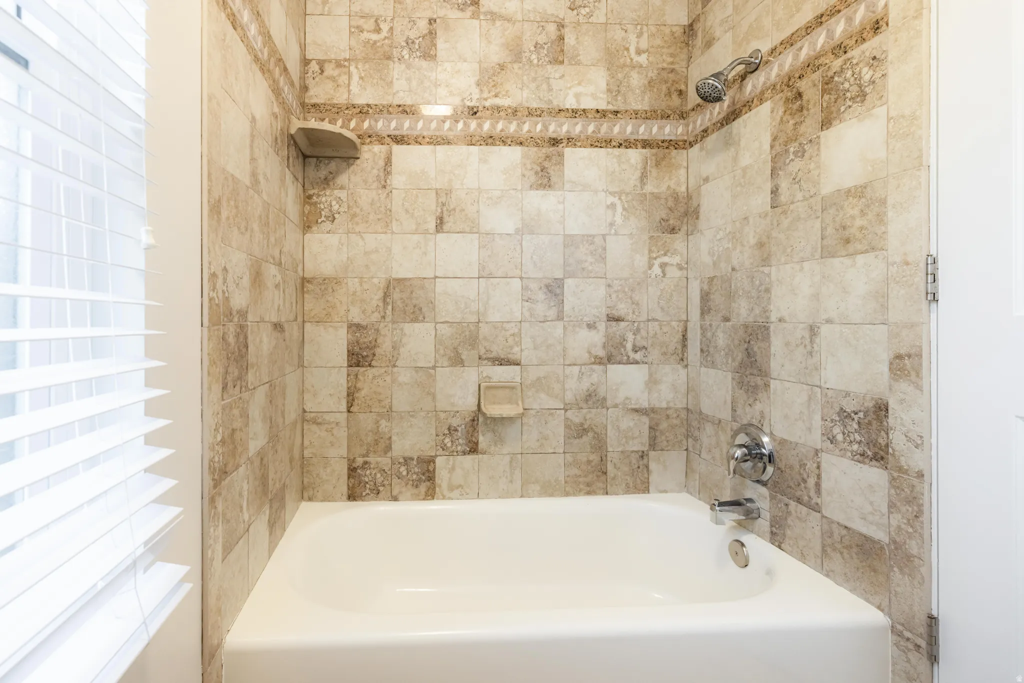 Bathtub with full-height natural stone-look tile surround, decorative accent band, integrated soap dish, and corner shelf