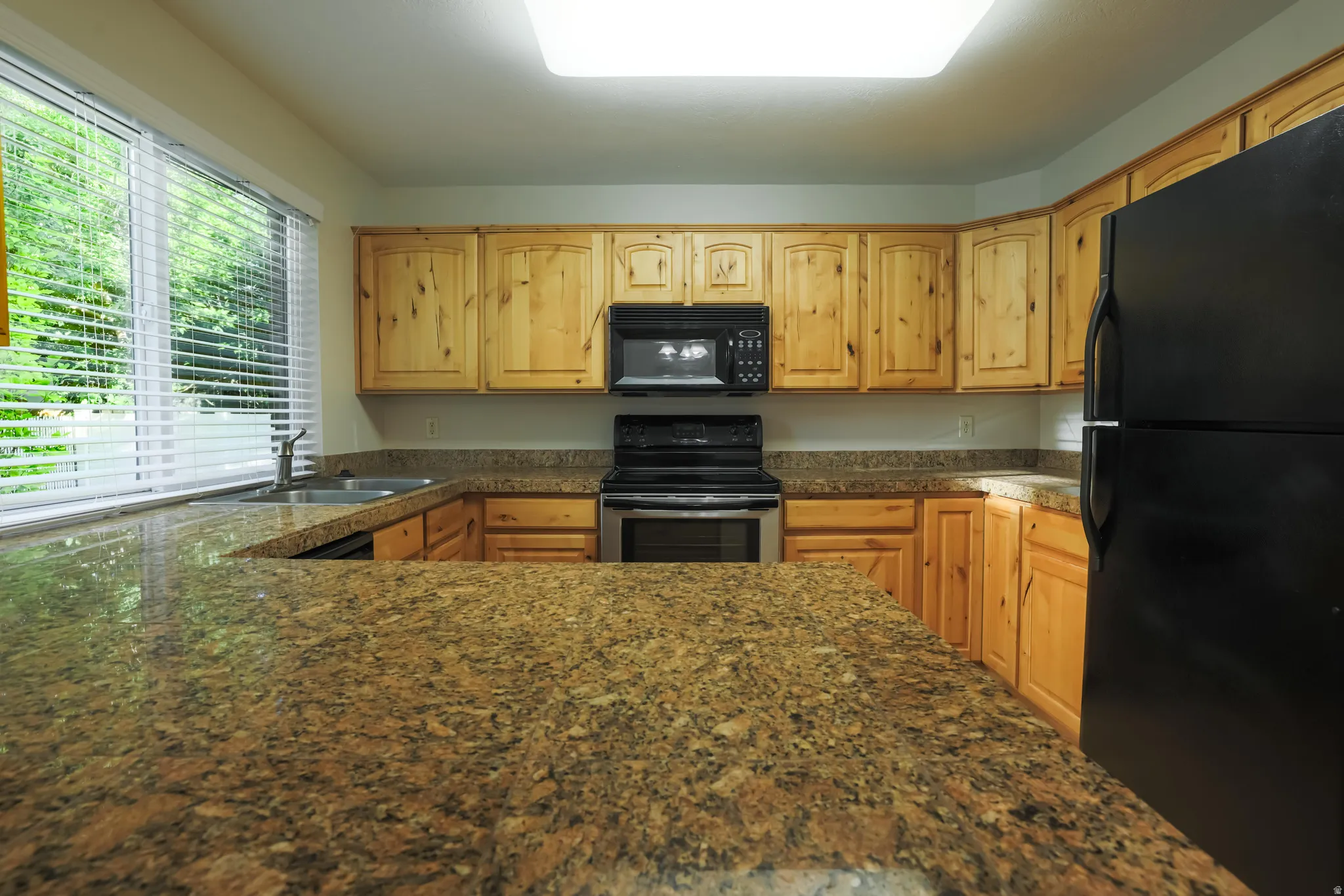 Kitchen featuring extensive stone countertops, natural wood cabinetry, stainless steel sink, black and stainless steel appliances, and a large window