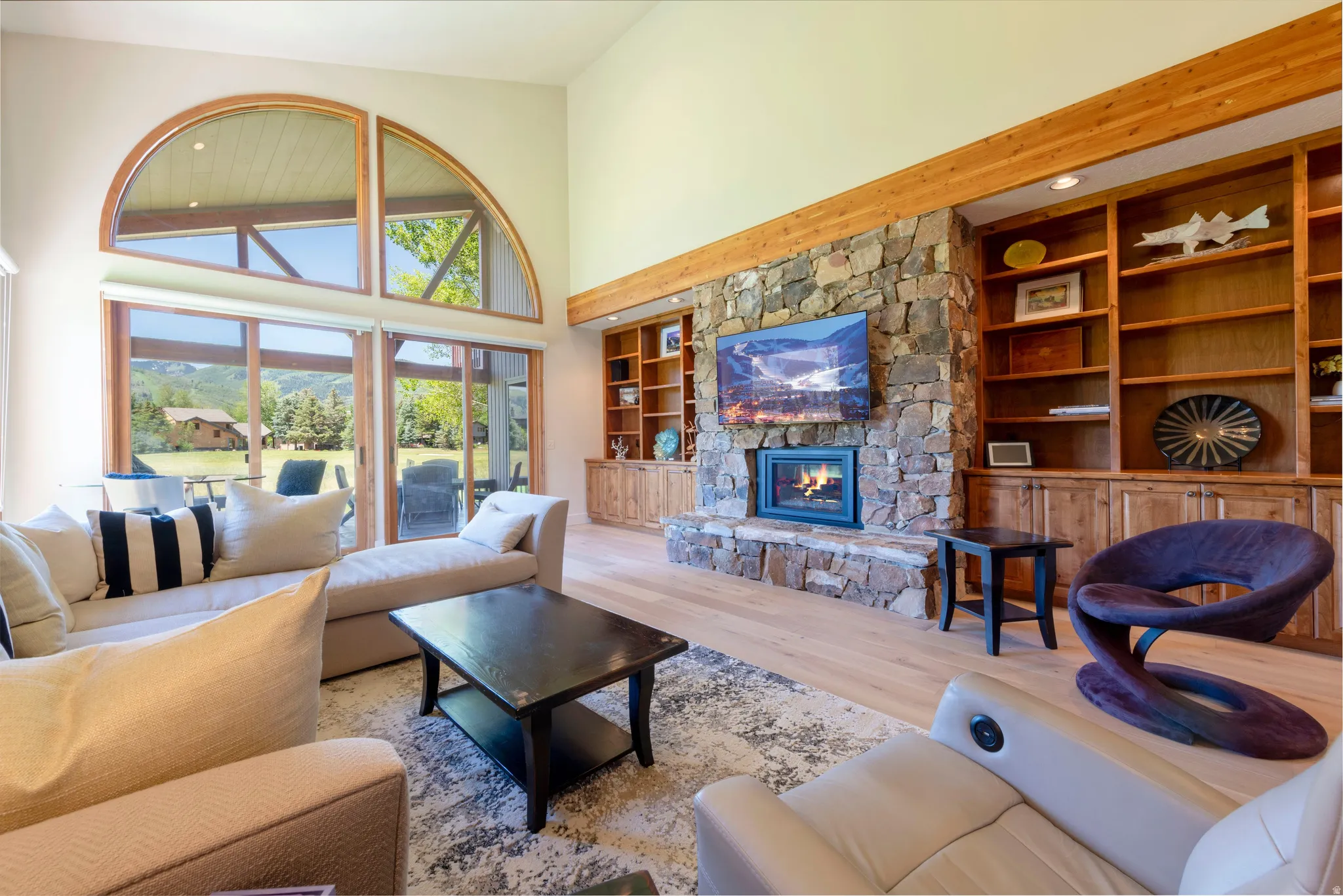 Spacious living area featuring a stone fireplace, built-in shelving, vaulted ceilings, light wood-finish flooring, and large windows with arched transoms