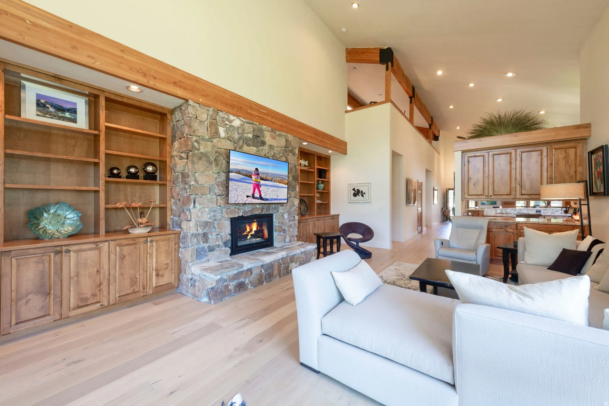 Spacious living area featuring a stone fireplace with hearth, built-in wooden shelving and cabinetry, high vaulted ceilings with exposed wood beams, and recessed lighting