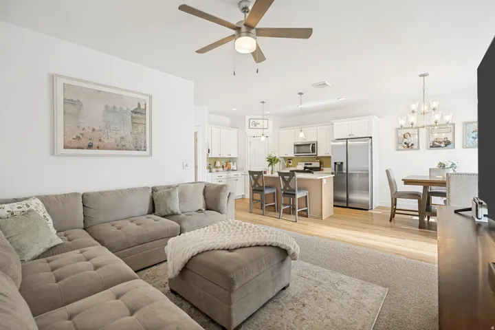 Open-concept living space featuring wood-finish flooring, a ceiling fan with integrated lighting, a kitchen island with pendant lighting, white cabinetry, and stainless steel appliances