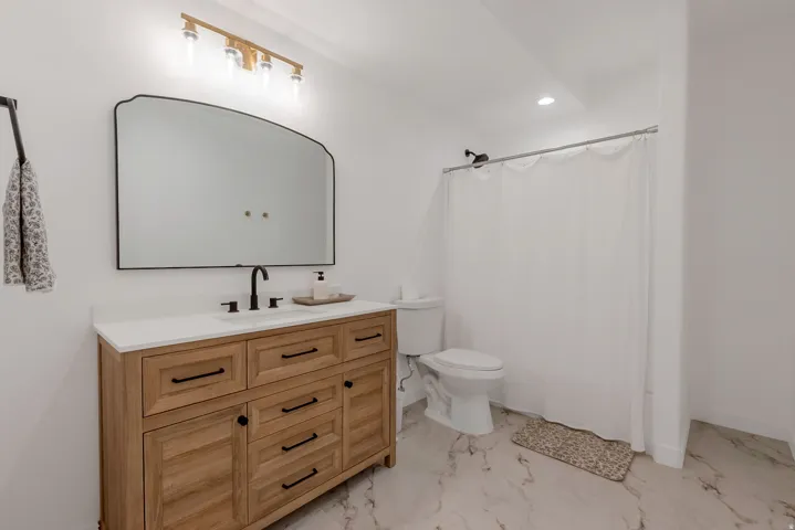 Spacious bathroom featuring a wood-finish vanity with a white countertop and matte black fixtures, an arched mirror, and a gold-tone wall-mounted light fixture